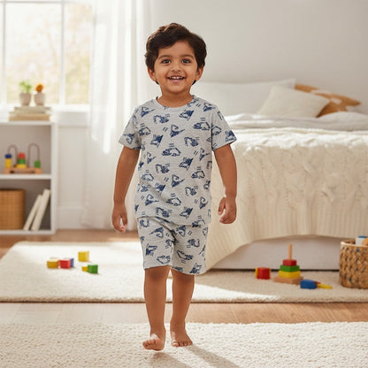 A smiling toddler boy wearing a grey kidshe coord set with blue construction vehicle prints in a bedroom.